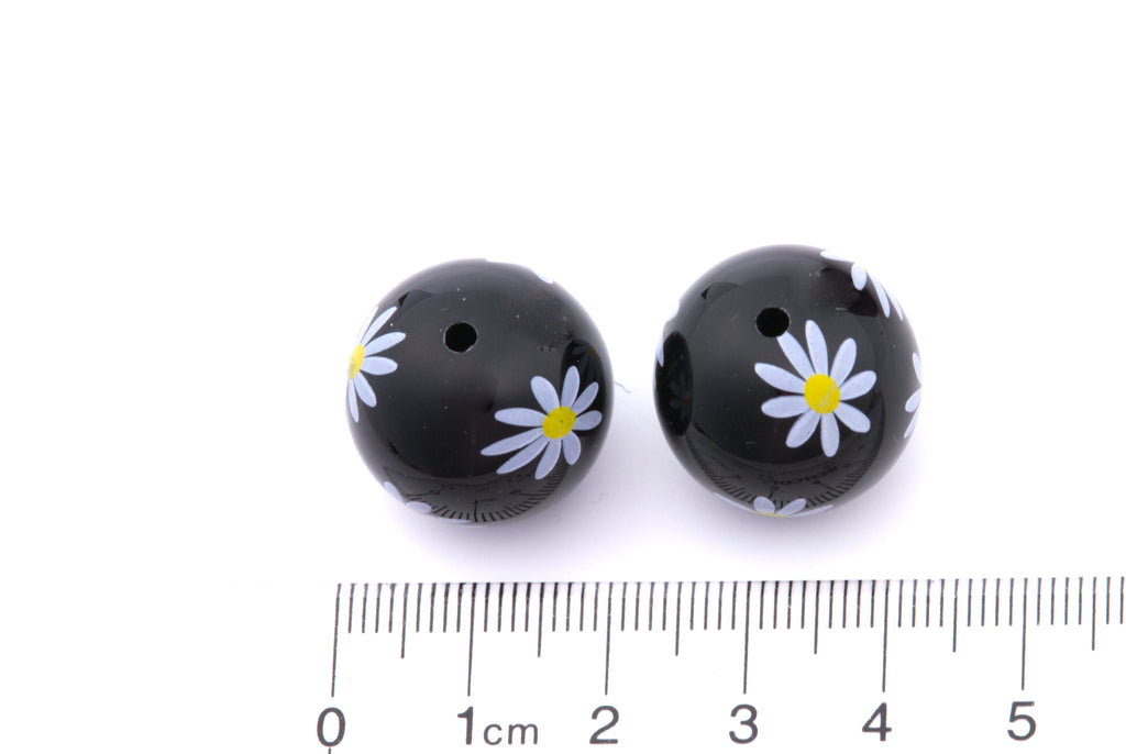 Two black beads with white flower designs on a white background, accompanied by a ruler for scale.