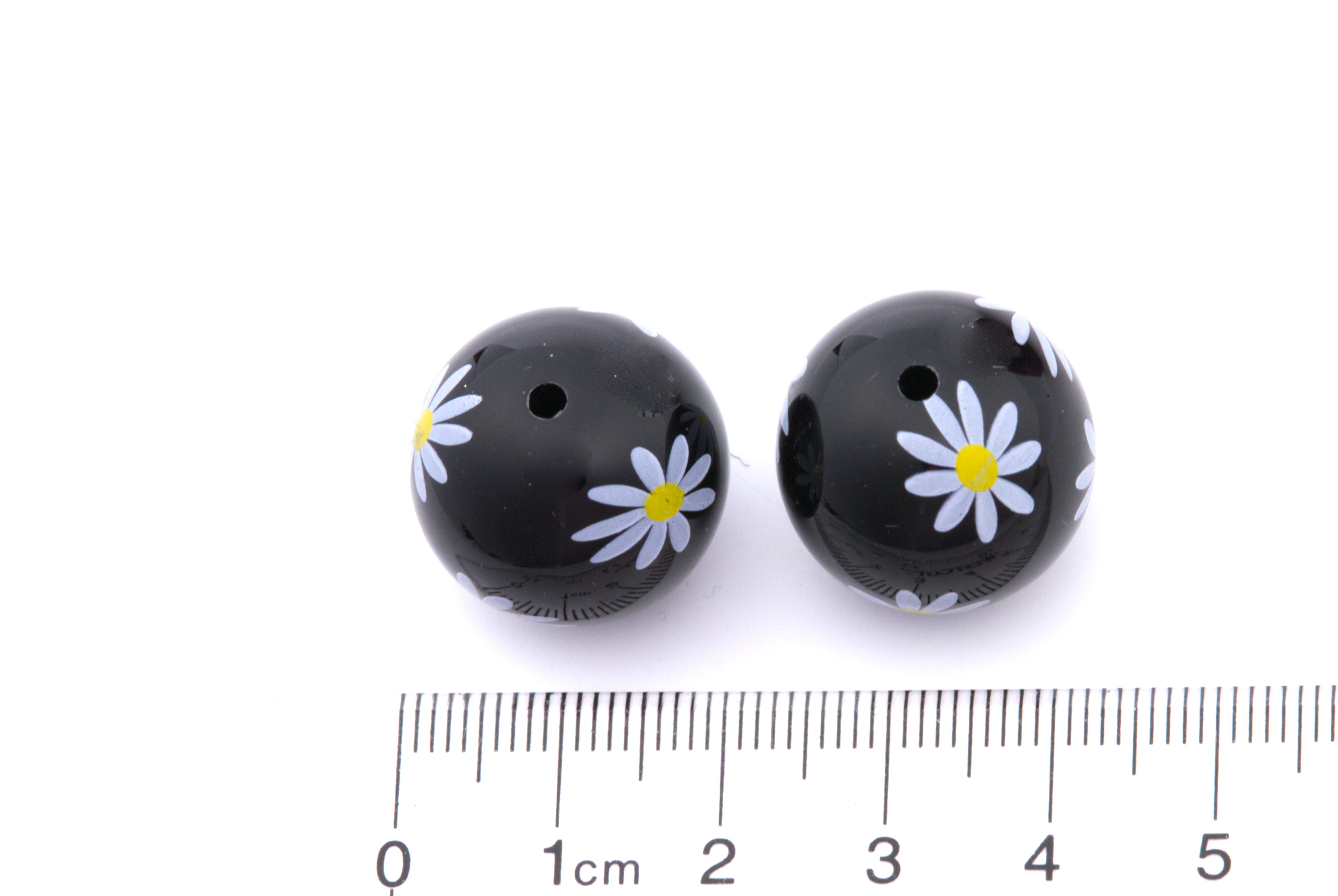 Two black beads with white flower designs on a white background, accompanied by a ruler for scale.