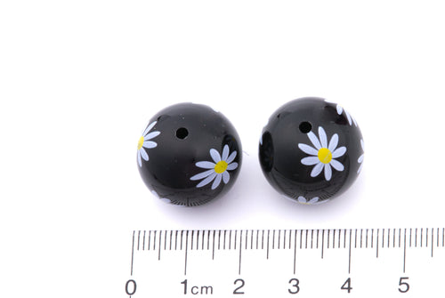 Two black beads with white flower designs on a white background, accompanied by a ruler for scale.