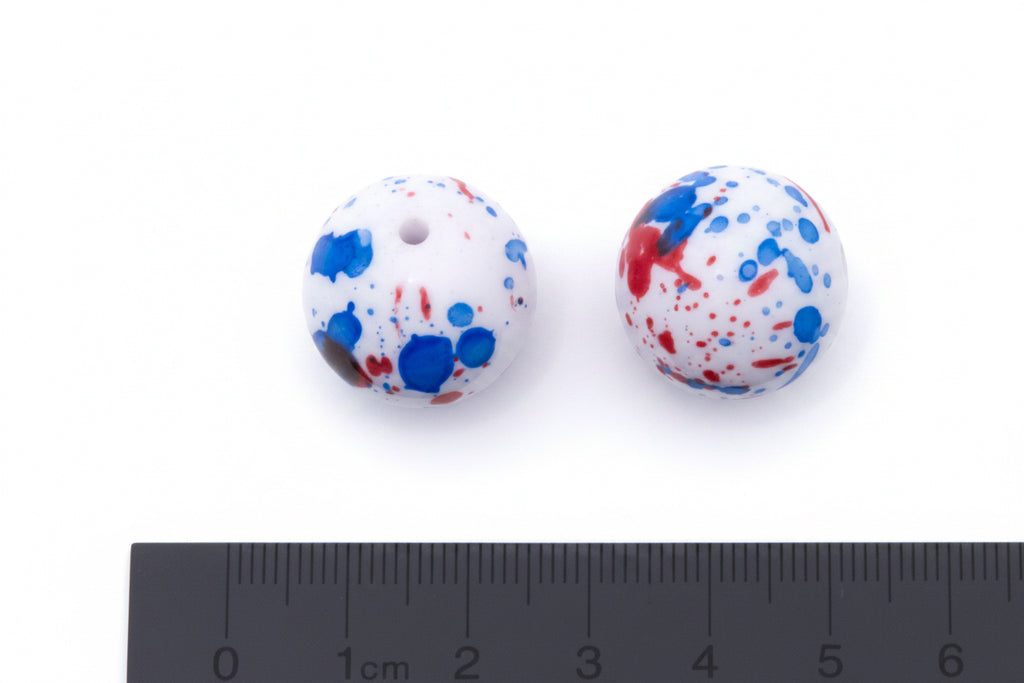 Two round white beads with blue and red paint splatter patterns, shown next to a black cm ruler for scale on a white background.