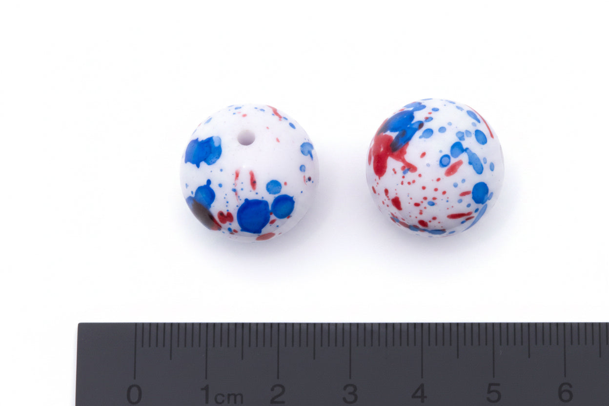 Two round white beads with blue and red paint splatter patterns, shown next to a black cm ruler for scale on a white background.