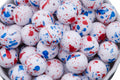White round beads with red and blue paint splatter patterns, shown in a close-up view inside a white container.
