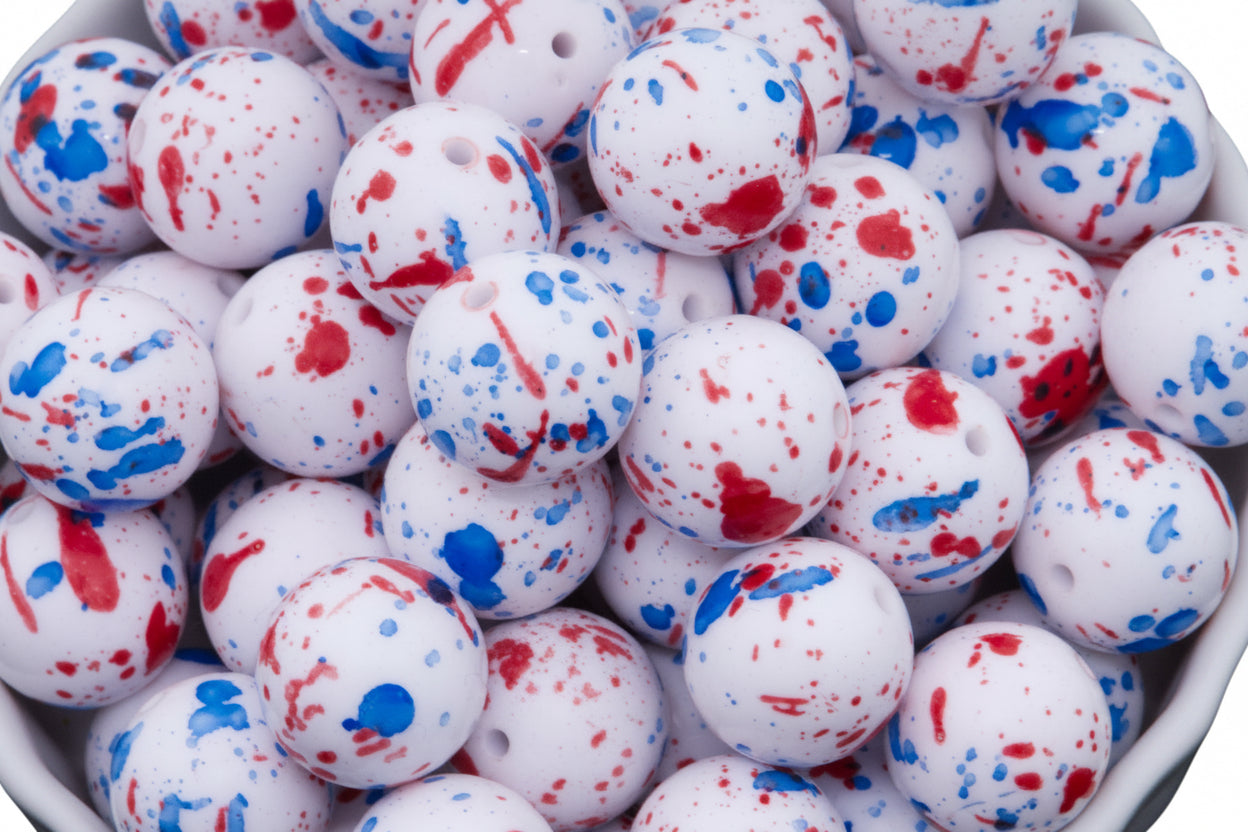 White round beads with red and blue paint splatter patterns, shown in a close-up view inside a white container.