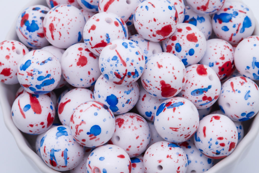 White round beads with red and blue paint splatter patterns, shown in a close-up view inside a white container.