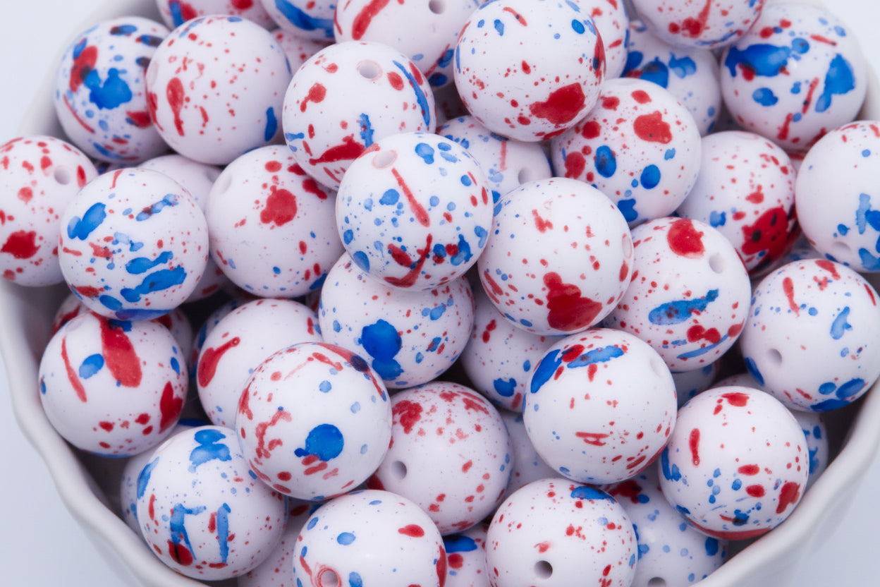 White round beads with red and blue paint splatter patterns, shown in a close-up view inside a white container.
