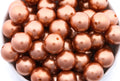 A close-up of shiny metallic copper brown beads piled in a white bowl, with the beads filling almost the entire frame.