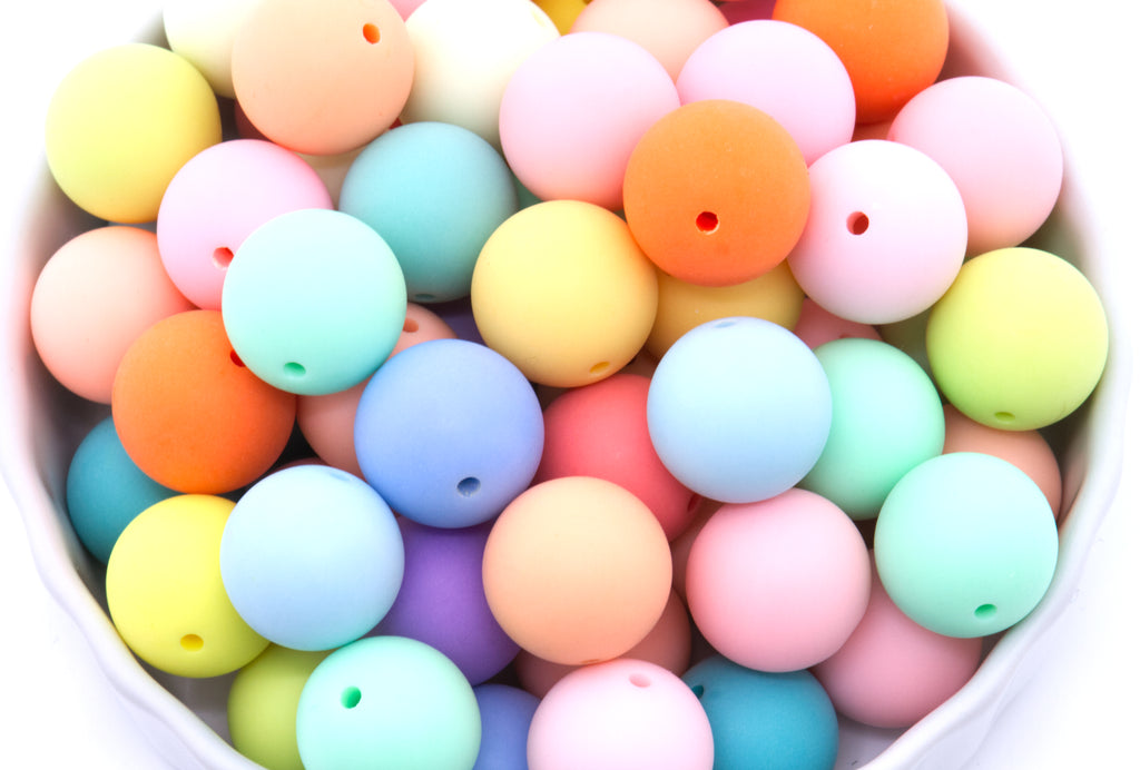 A close-up of round frosted matte rainbow beads in multiple colors, including pink, blue, yellow, orange, mint green, and purple, piled inside a white bowl.