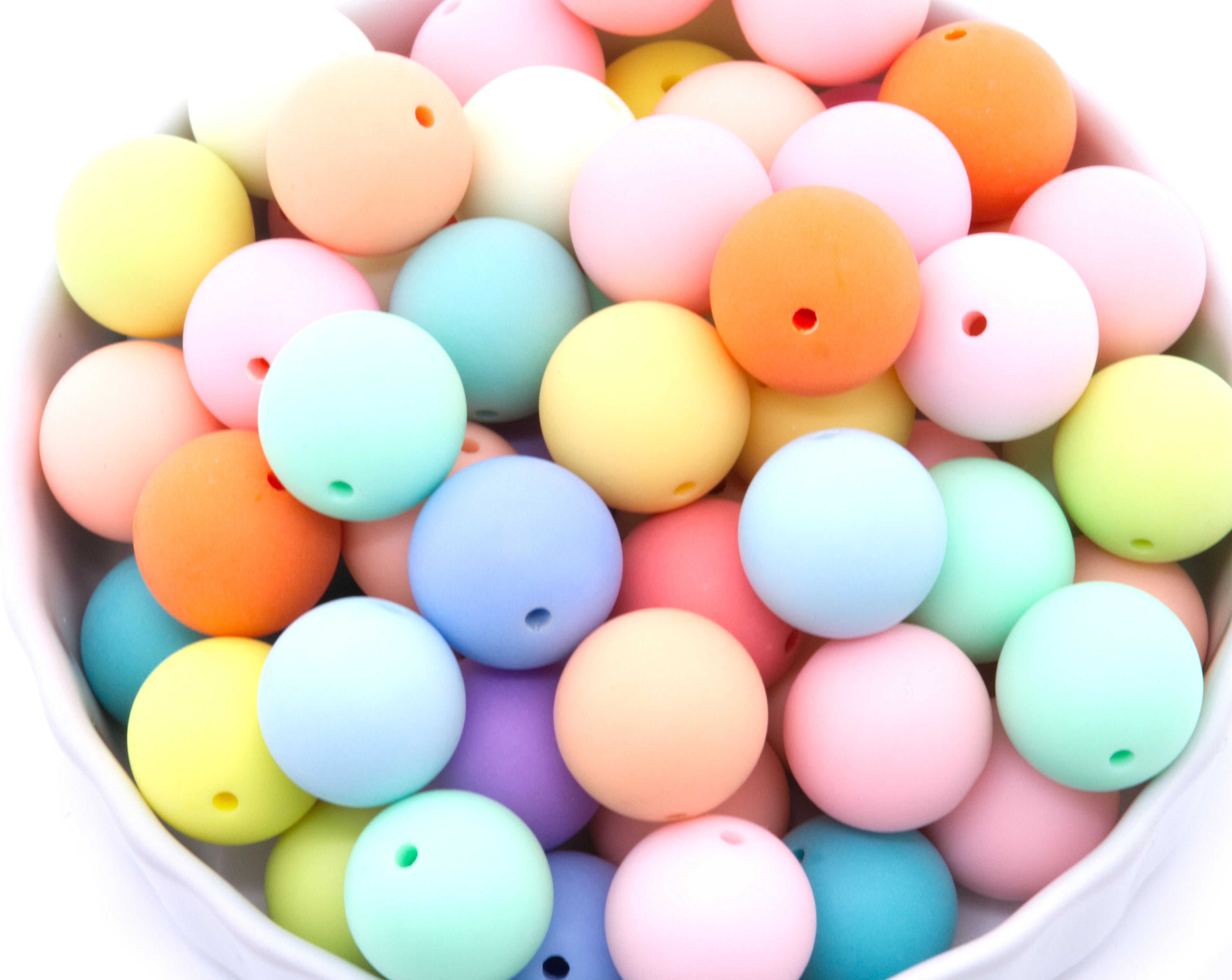 A close-up of round frosted matte rainbow beads in multiple colors, including pink, blue, yellow, orange, mint green, and purple, piled inside a white bowl.