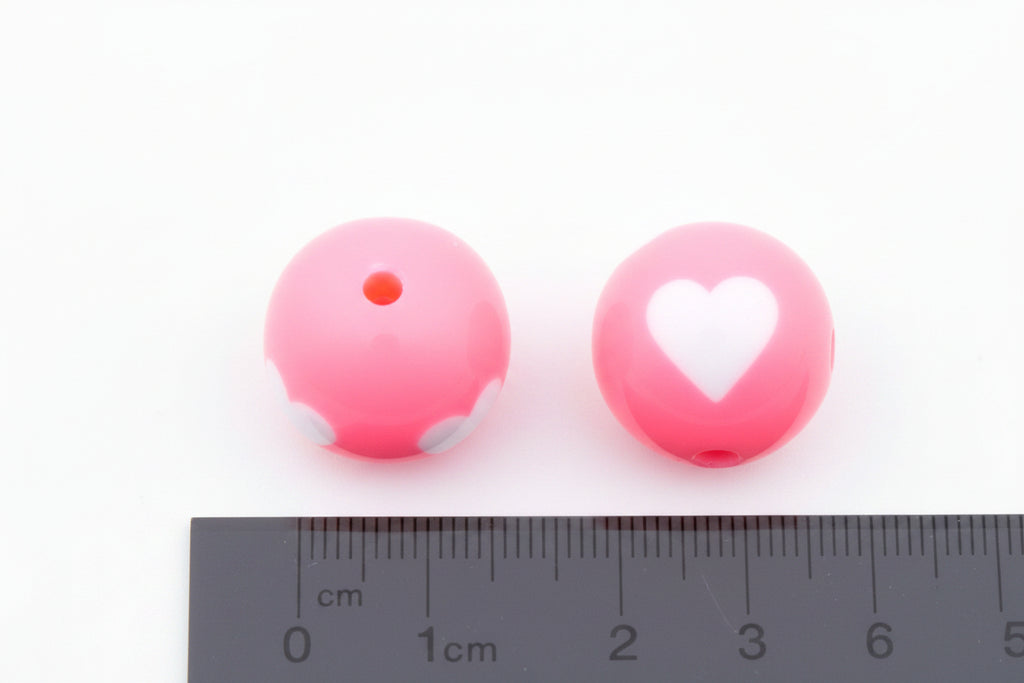 Two pink beads with a heart design and one plain, next to a ruler for scale on a white background.