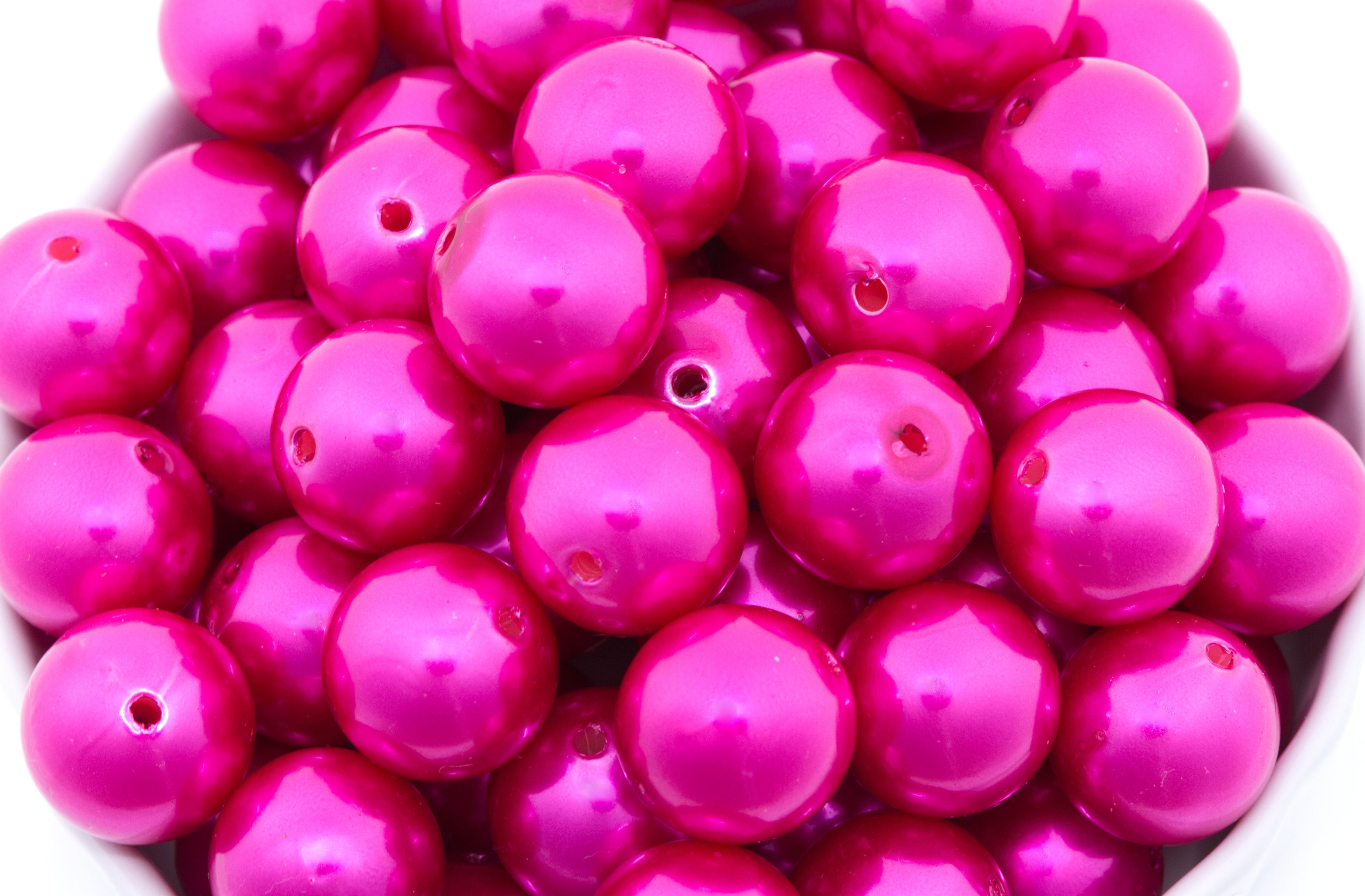 A close-up pile of glossy, metallic magenta pink round beads with pre-drilled holes, filling the frame against a white background.