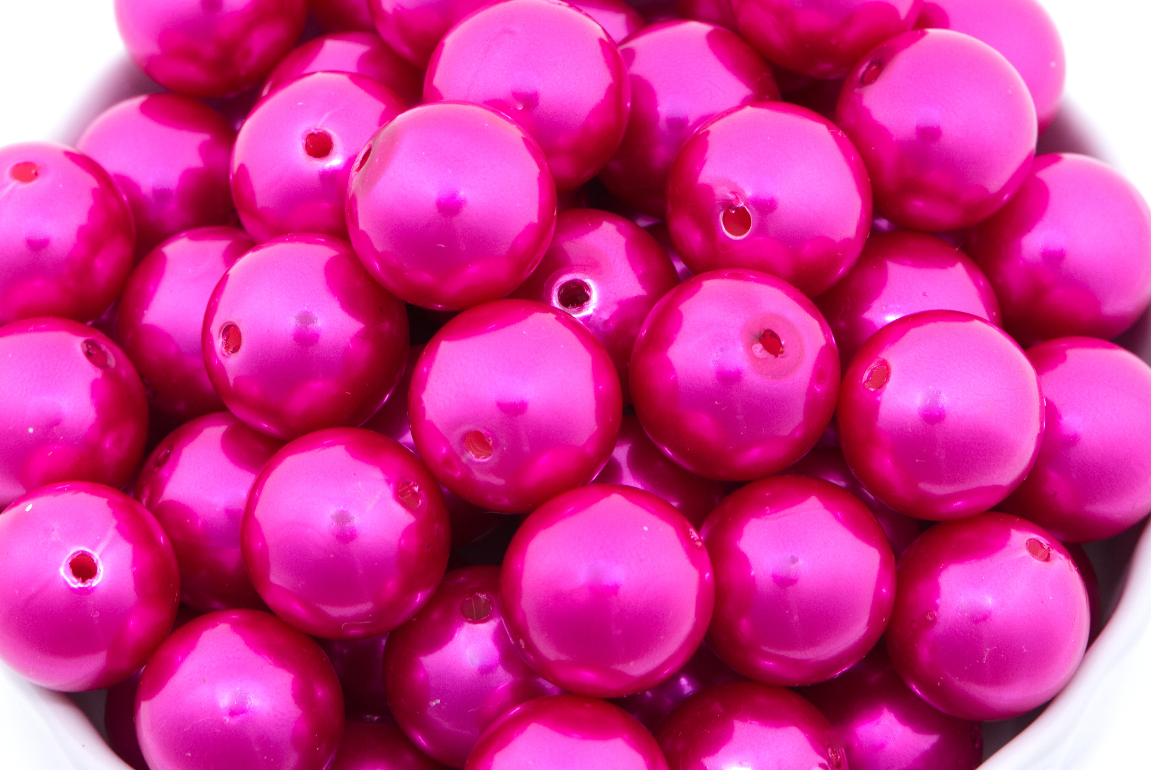 A close-up of glossy hot pink round beads with small holes, piled together in a white bowl.