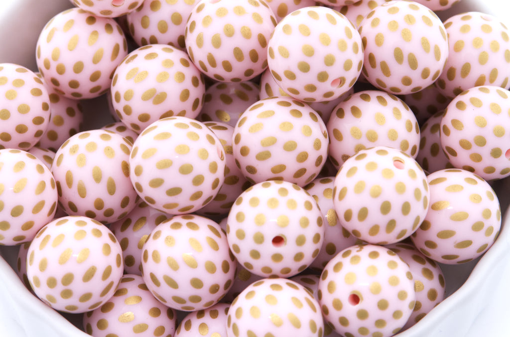 A close-up of round light pink beads with metallic gold polka dots, piled inside a white container.