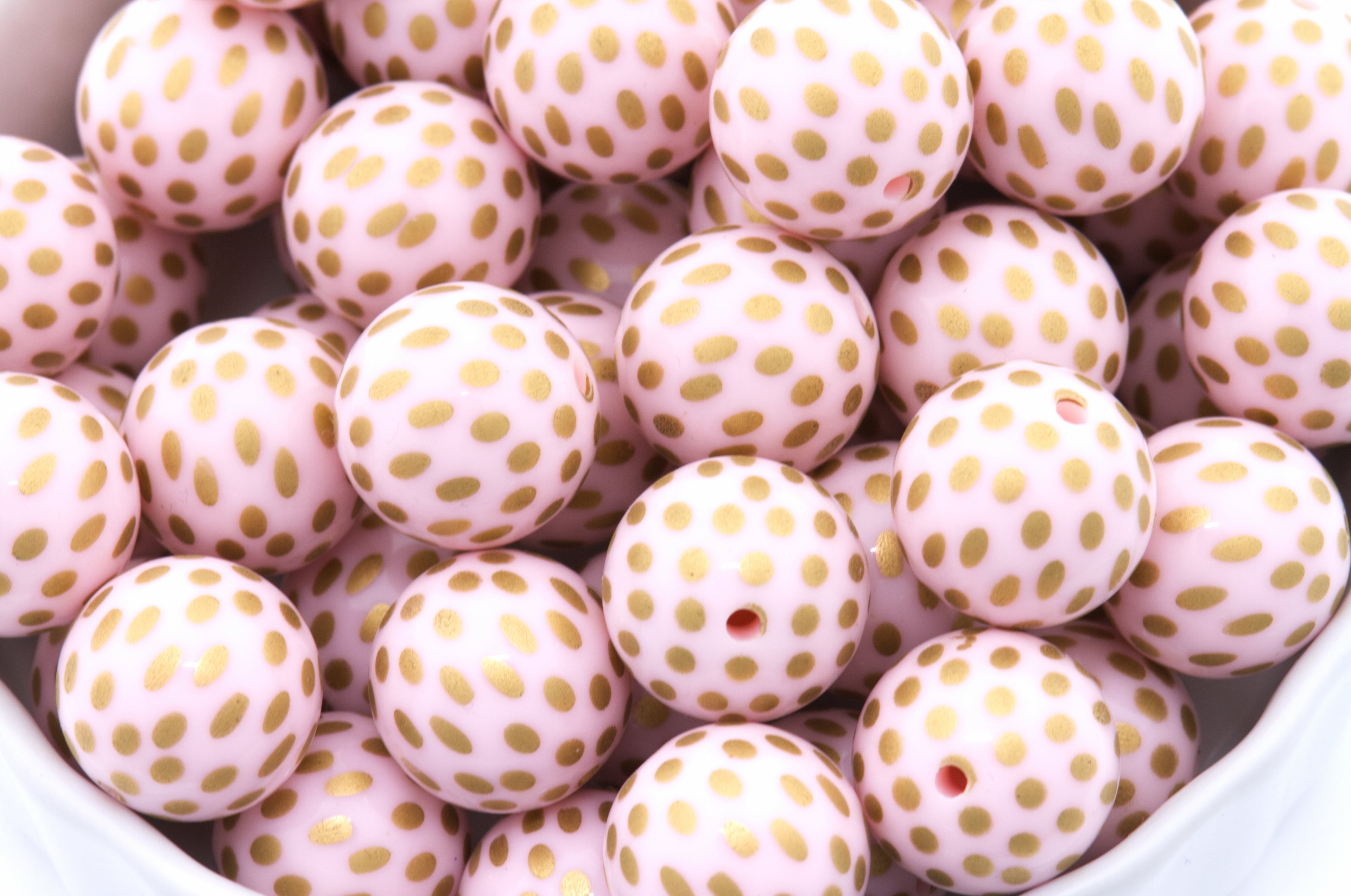 A close-up of round light pink beads with metallic gold polka dots, piled inside a white container.