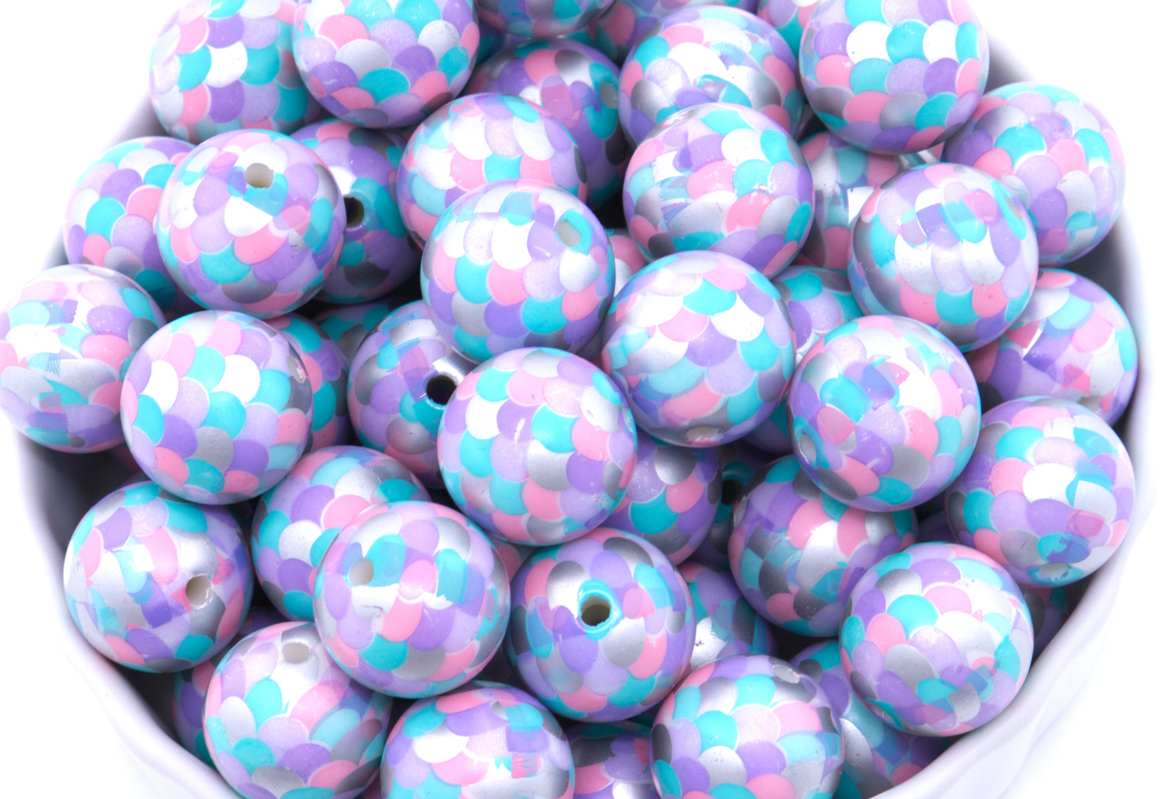 A pile of round, purple, pink and blue mermaid scale print beads, arranged in a white bowl against a white background.