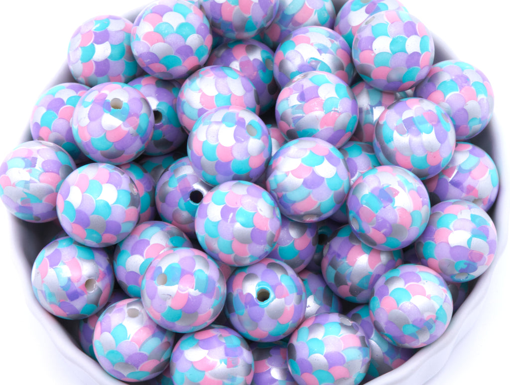 A pile of round, purple, pink and blue mermaid scale print beads, arranged in a white bowl against a white background.