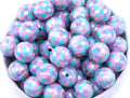 A pile of round, purple, pink and blue mermaid scale print beads, arranged in a white bowl against a white background.
