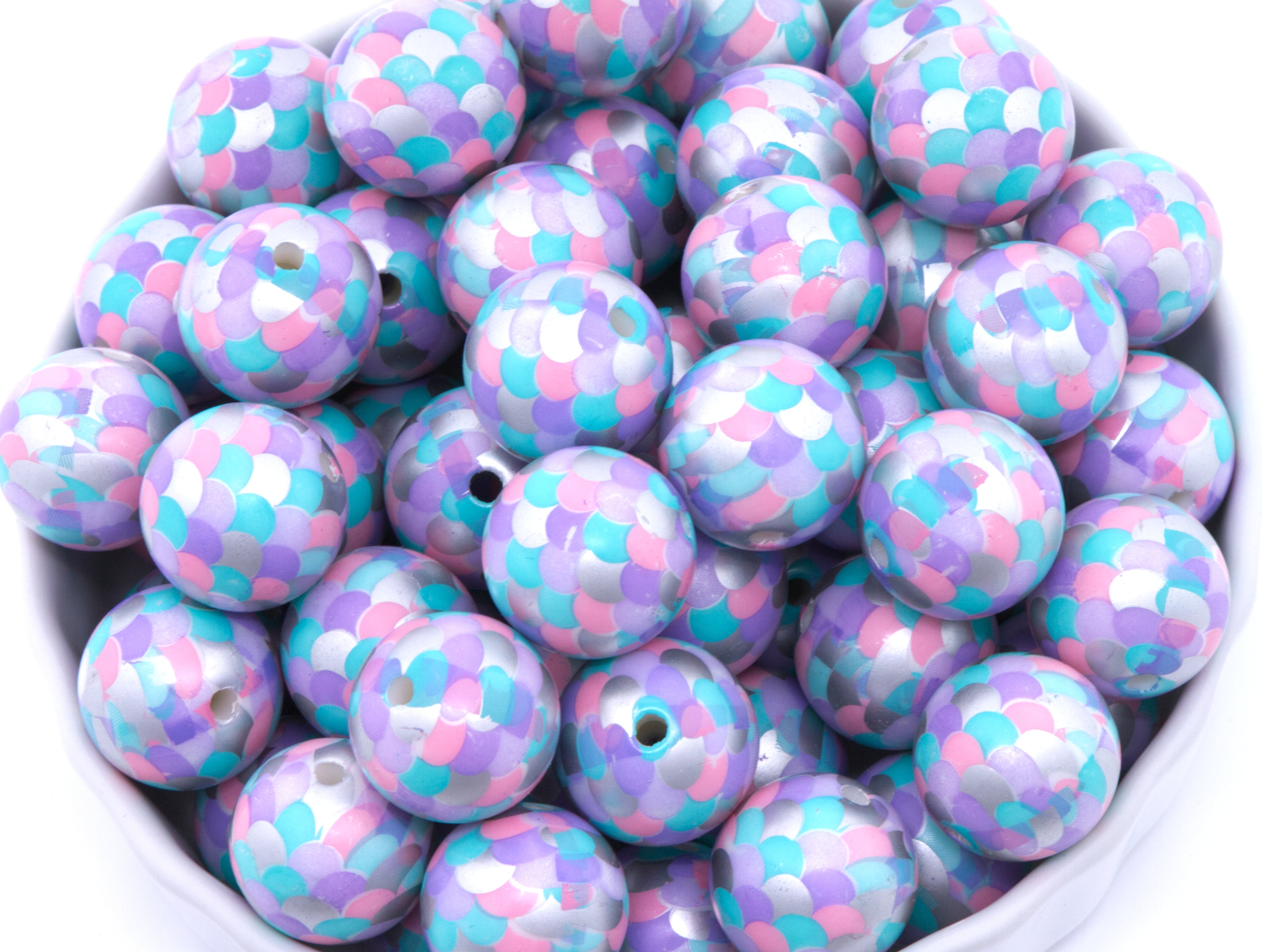 A pile of round, purple, pink and blue mermaid scale print beads, arranged in a white bowl against a white background.