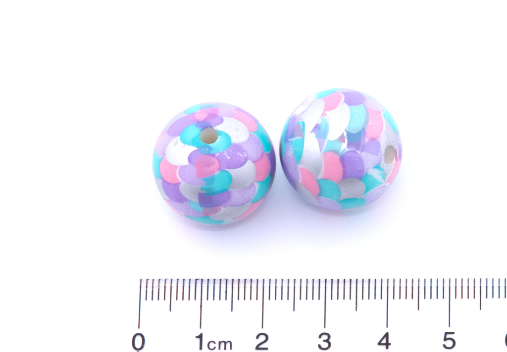 Two round, purple, pink and blue mermaid scale print beads next to a cm ruler for scale on a white background.