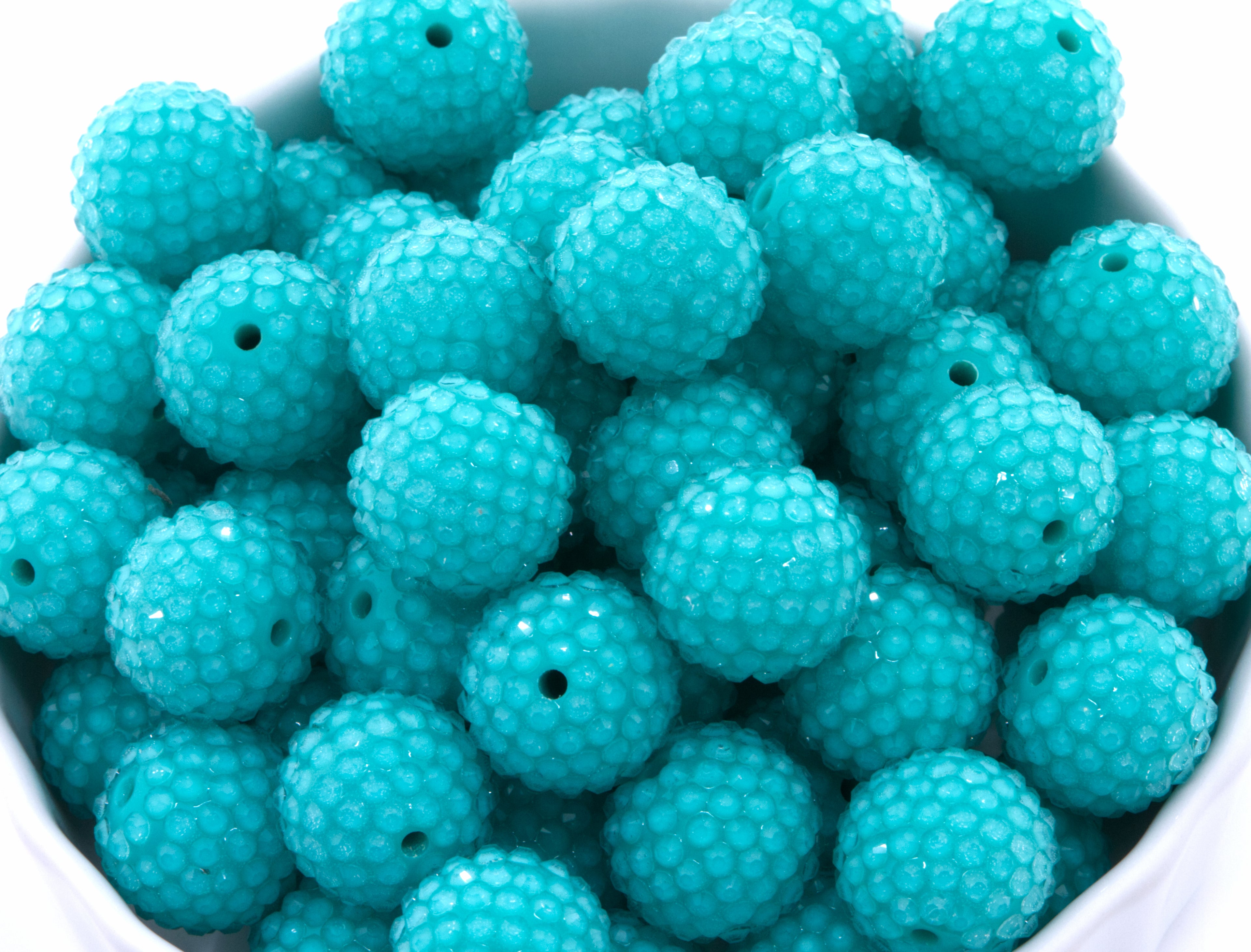 A close-up of teal blue rhinestone bubblegum beads, piled inside a white container on a white background. 