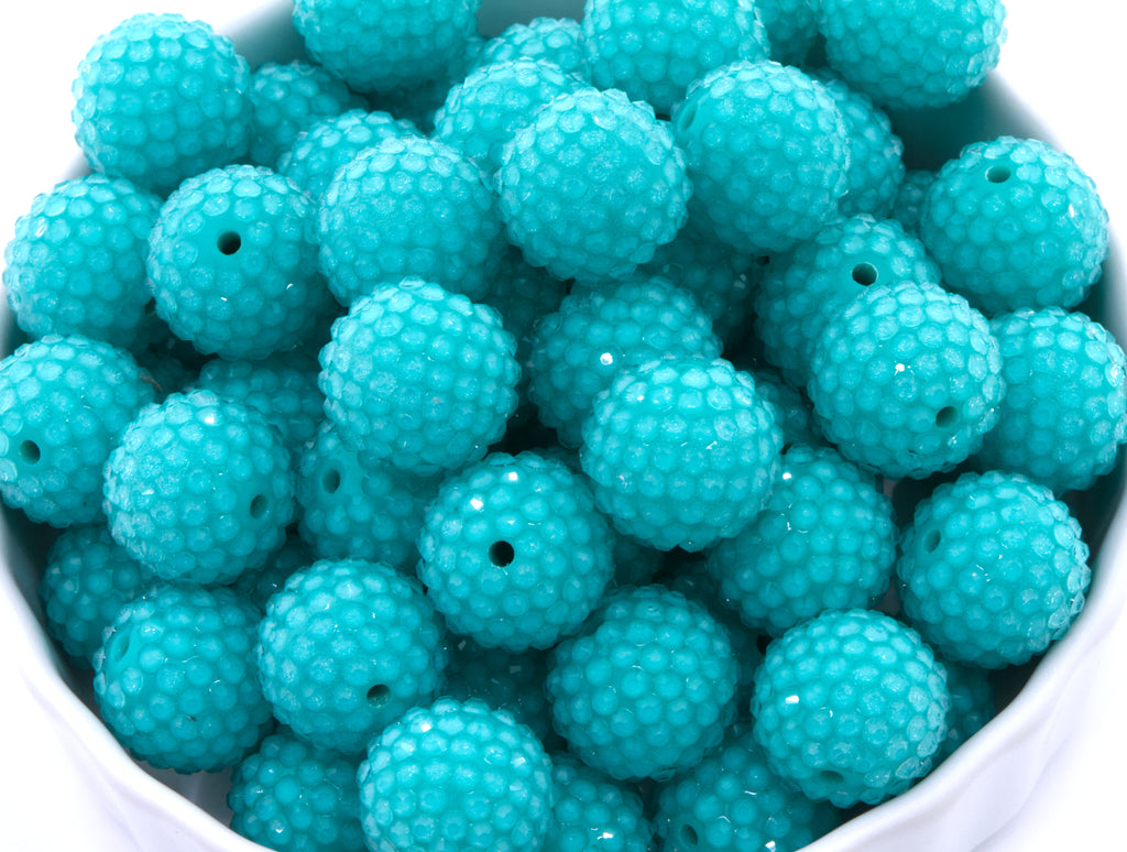 A close-up of teal blue rhinestone bubblegum beads, piled inside a white container on a white background. 