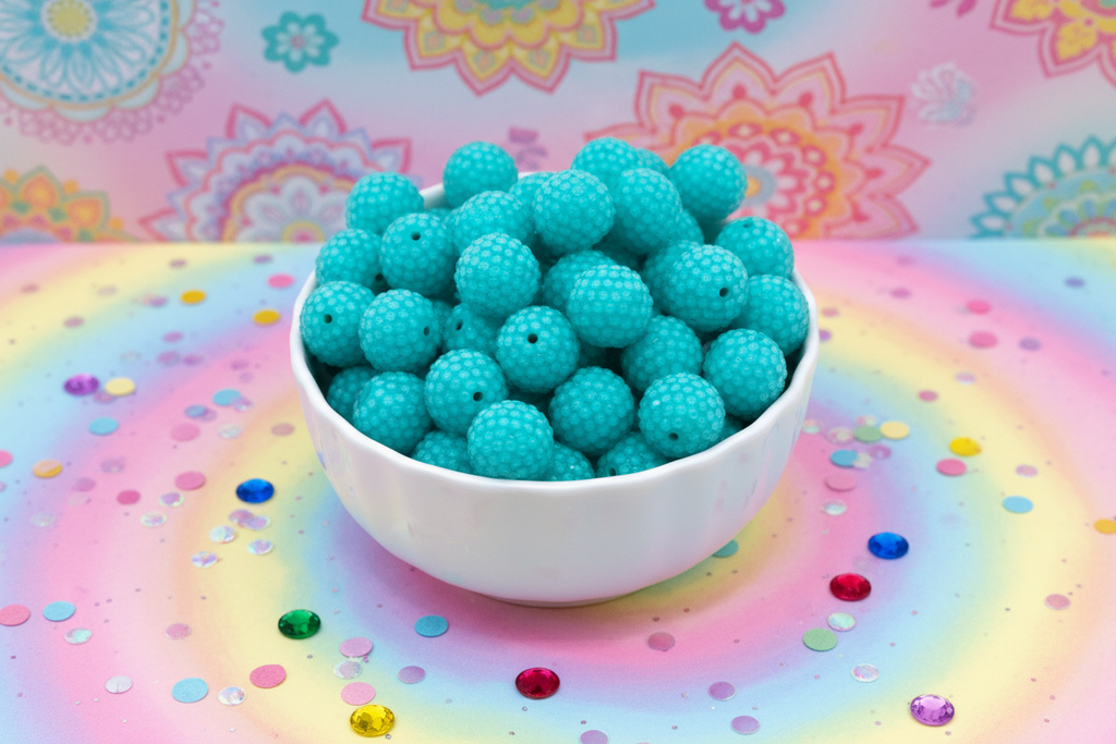 A white ceramic bowl filled with teal blue rhinestone bubblegum beads, set against a pastel rainbow background with colorful confetti and floral mandala patterns.