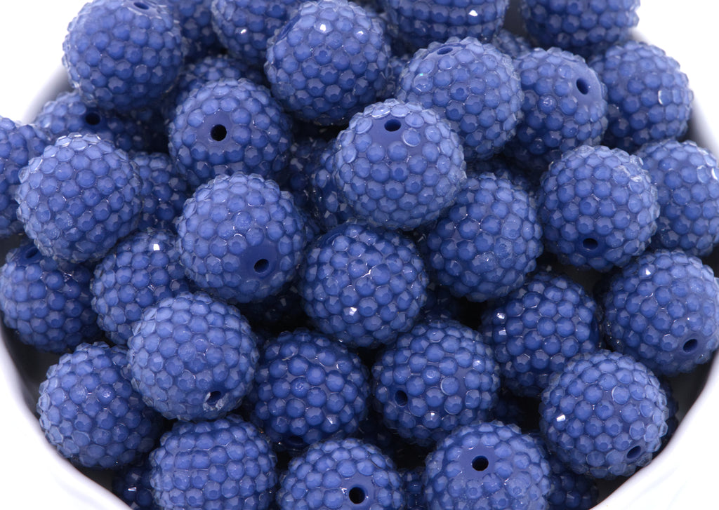 A close-up of many blue rhinestone beads piled together in a white bowl on a white background. 