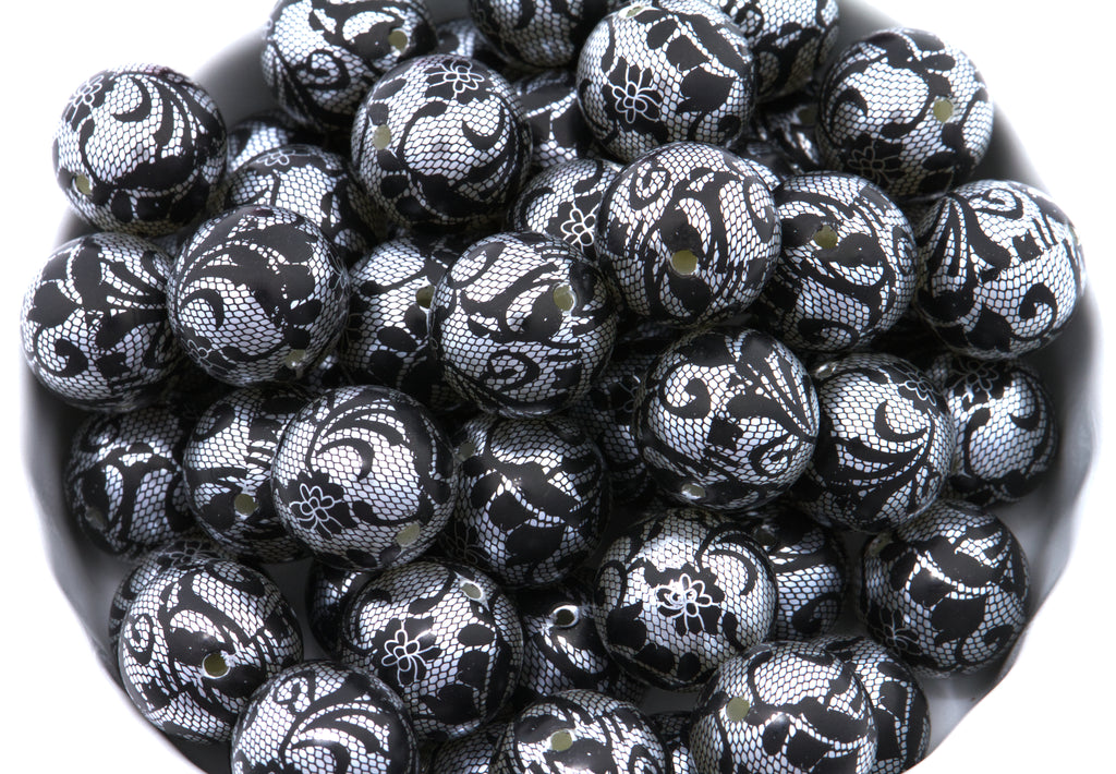 A pile of round black and silver lace patterned beads , shown in a close-up view inside a white container.