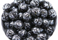A pile of round black and silver lace patterned beads , shown in a close-up view inside a white container.