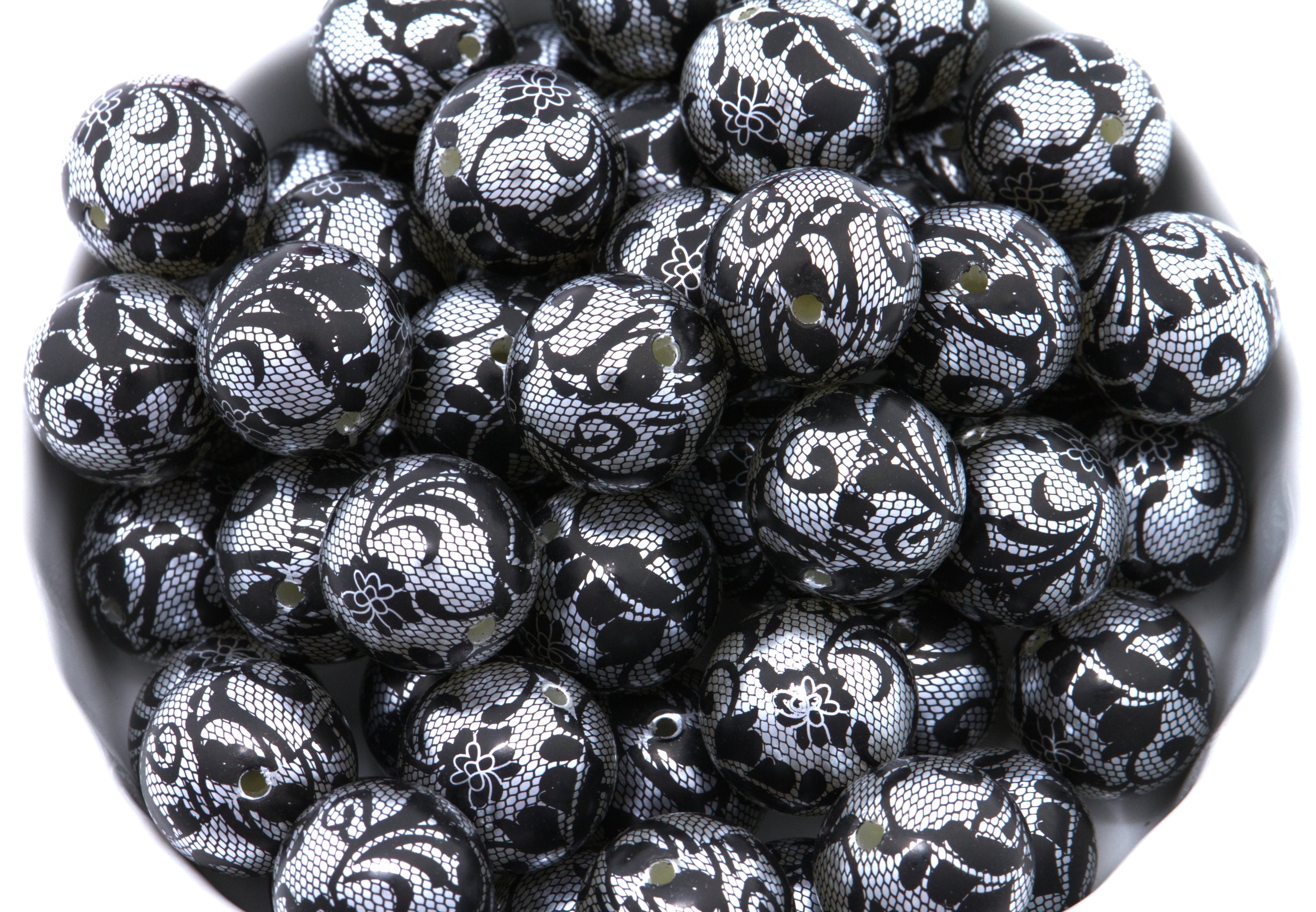 A pile of round black and silver lace patterned beads , shown in a close-up view inside a white container.