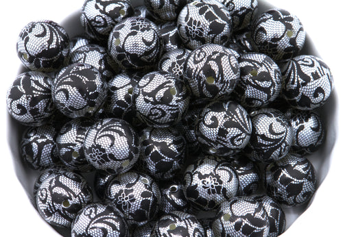 A pile of round black and silver lace patterned beads , shown in a close-up view inside a white container.