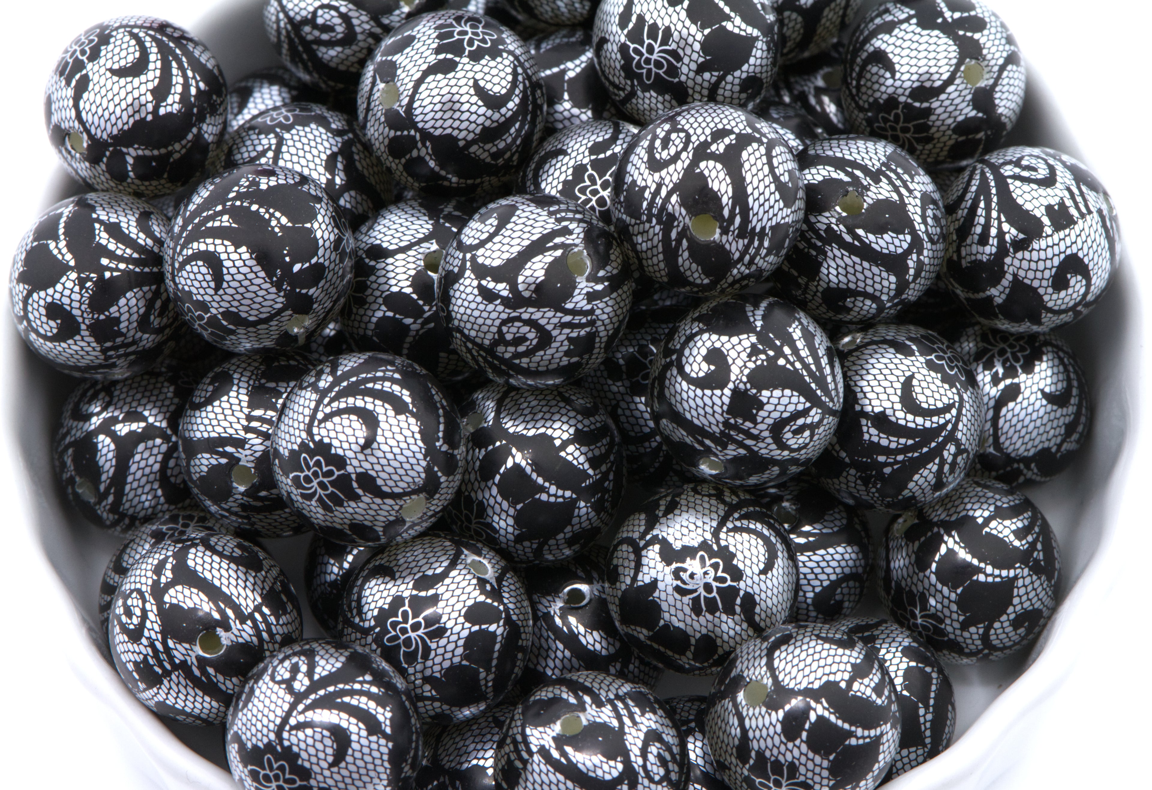 A pile of round black and silver lace patterned beads , shown in a close-up view inside a white container.