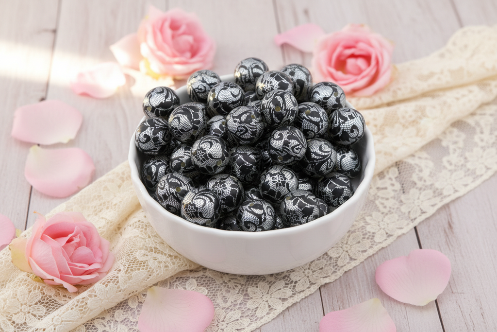 A white ceramic bowl filled with black and silver lace patterned round beads, styled on a light wood surface with pink roses, lace fabric, and scattered pink petals.