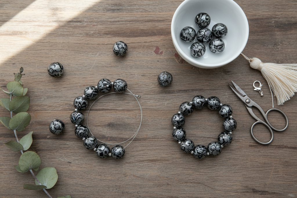 Black and silver lace patterned beads arranged on a wooden surface, with two wire loops holding bead strands, a small white bowl of loose beads, silver scissors, a cream tassel, and eucalyptus leaves.