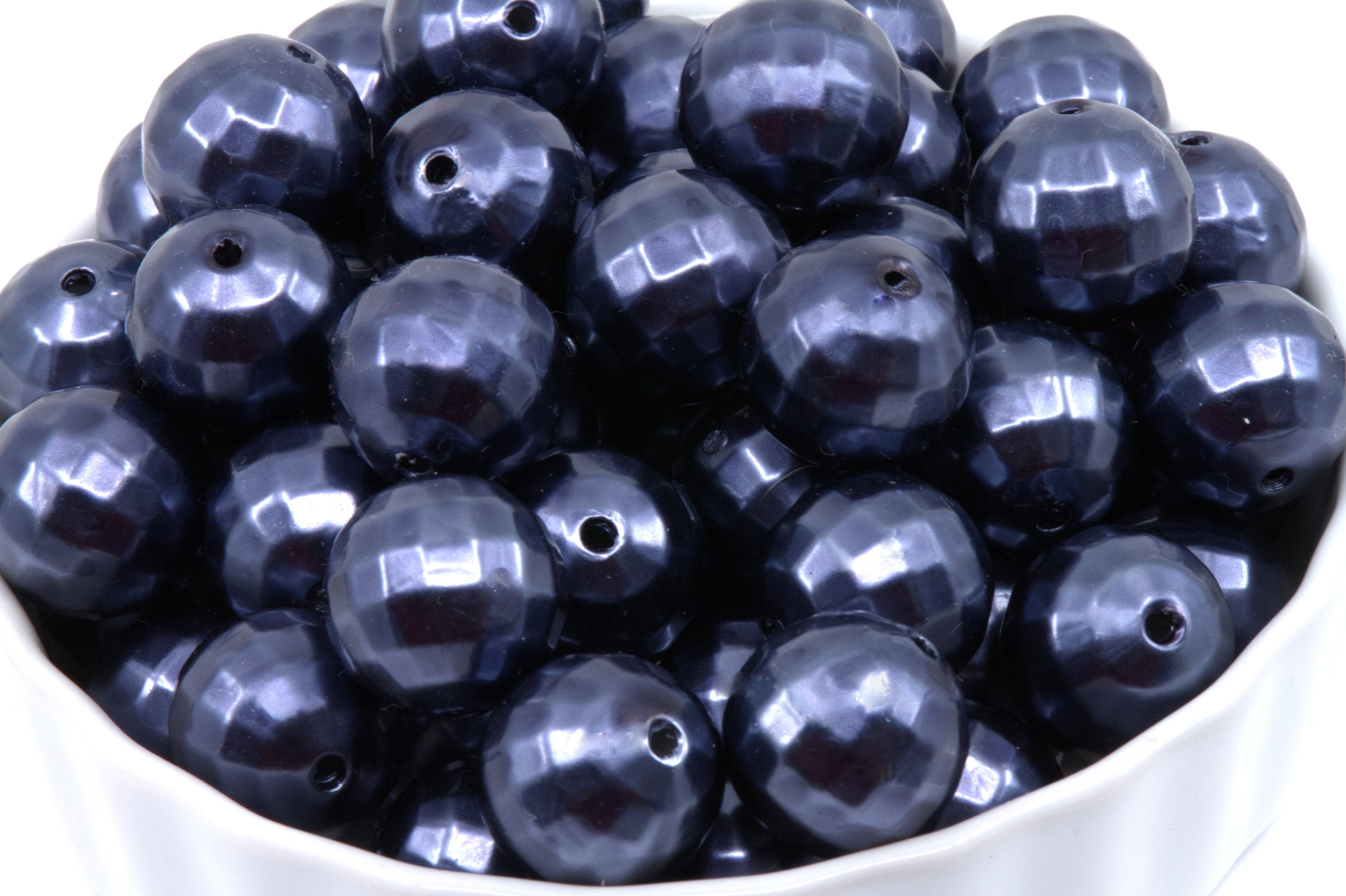 A pile of faceted dark blue bubblegum beads displayed in a white bowl against a white background.