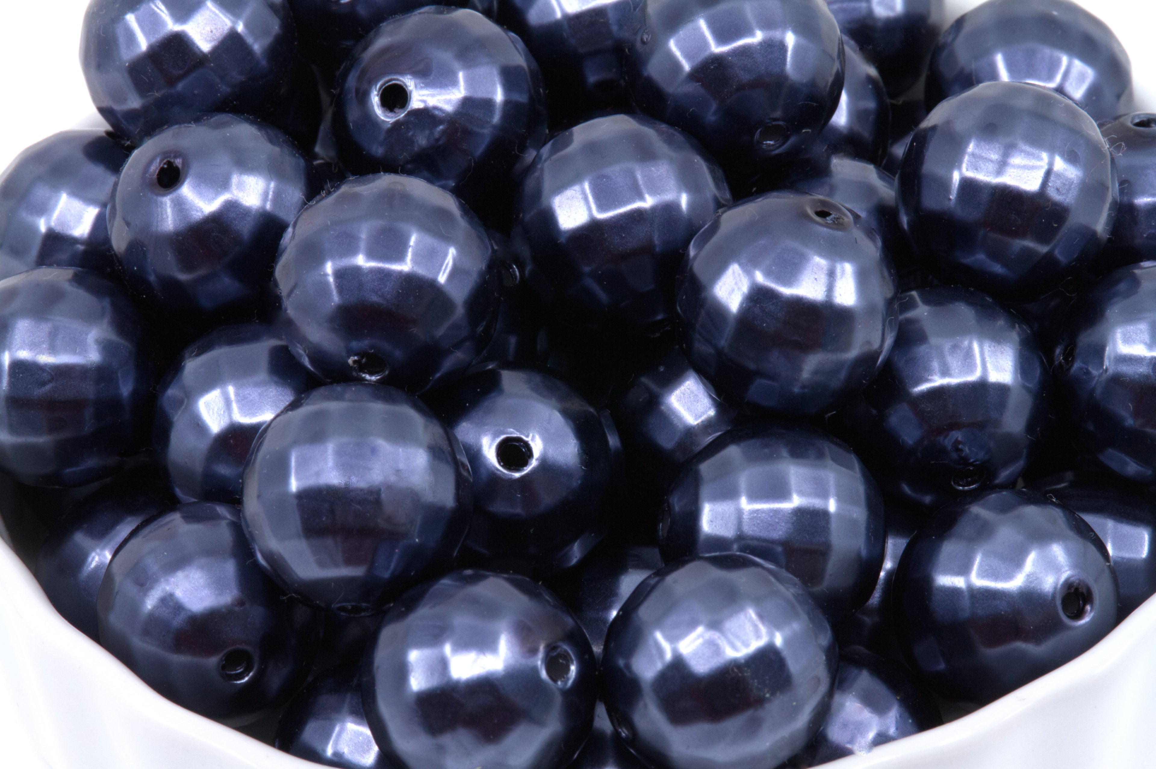 A pile of faceted dark blue bubblegum beads displayed in a white bowl against a white background.