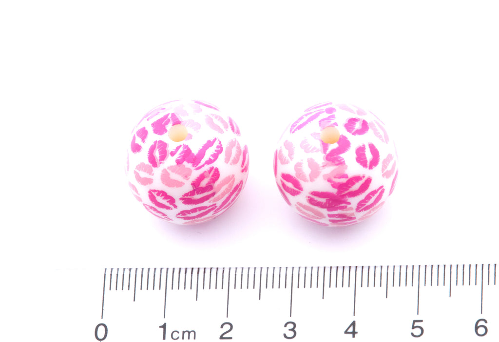 Two pink and white kiss patterned beads with a ruler for scale on a white background