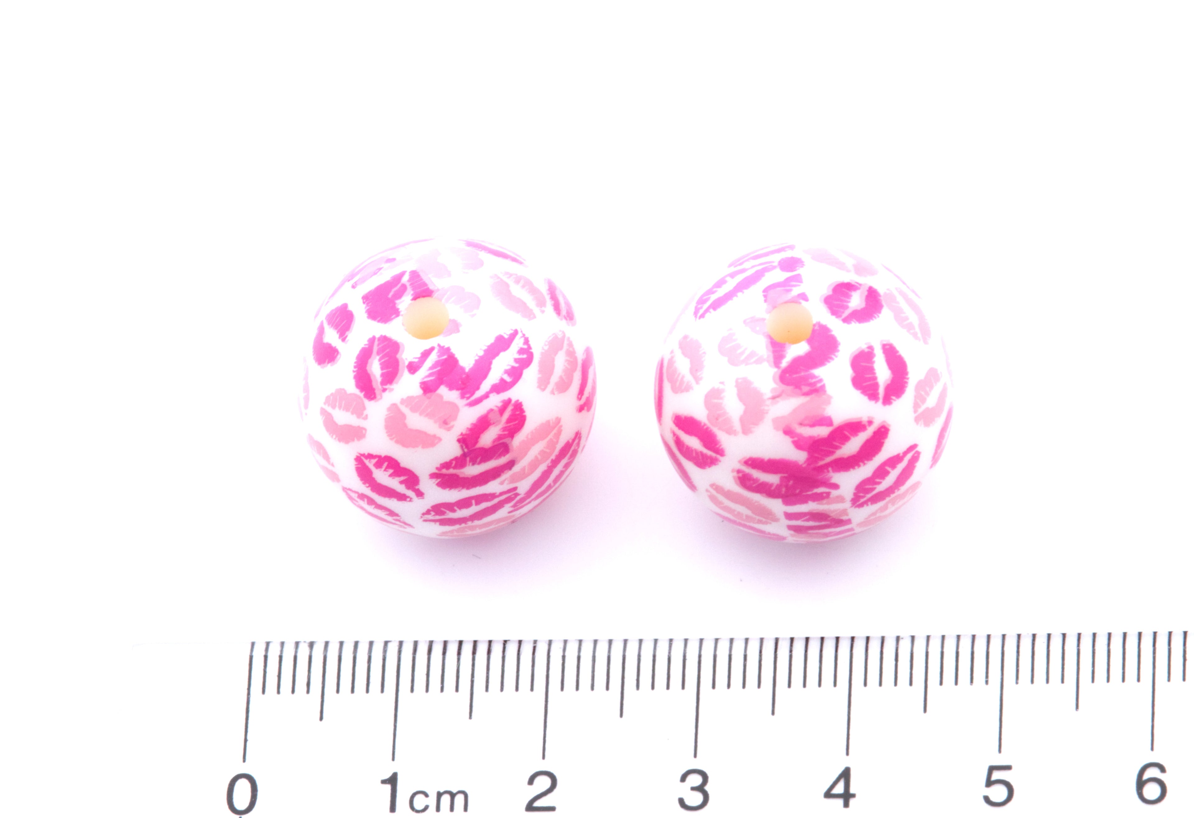 Two pink and white kiss patterned beads with a ruler for scale on a white background