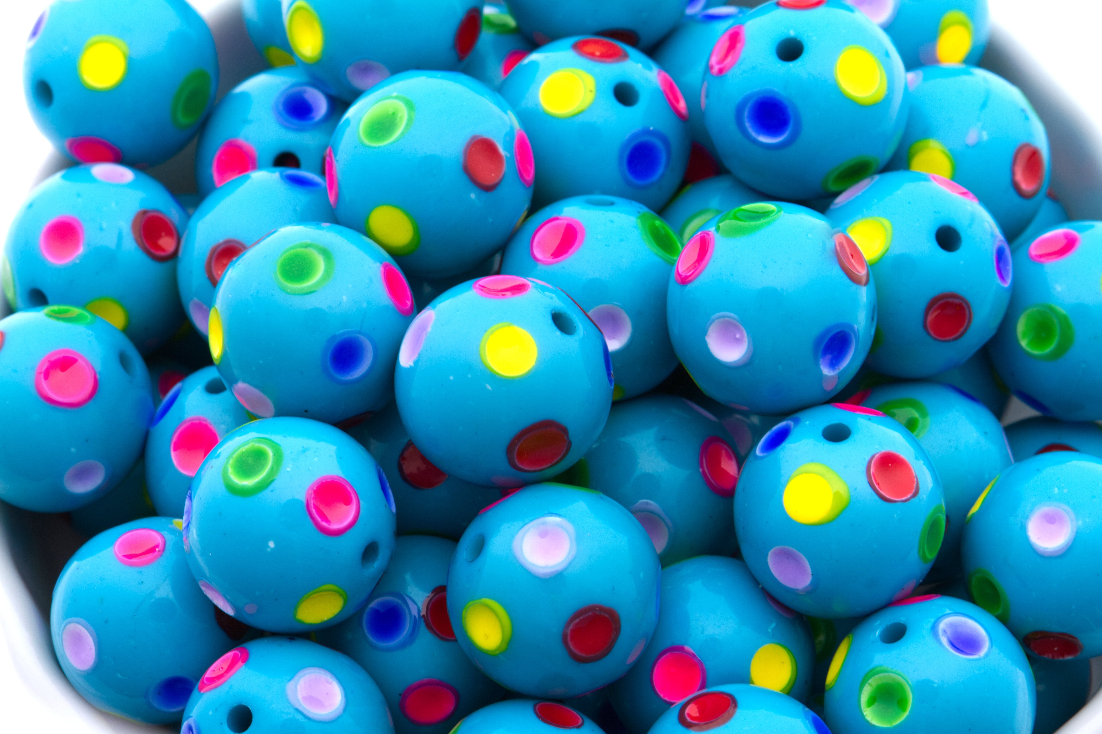 A close-up of many bright blue round beads with small multicolored polka dots, piled inside a white bowl against a white background.