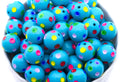 A close-up of many bright blue round beads with small multicolored polka dots, piled inside a white bowl against a white background.