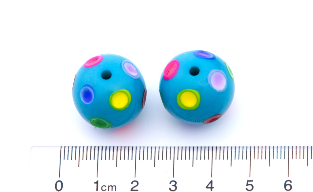 Two small blue round beads with colorful printed polka dots, shown next to a centimeter ruler for scale on a white background.