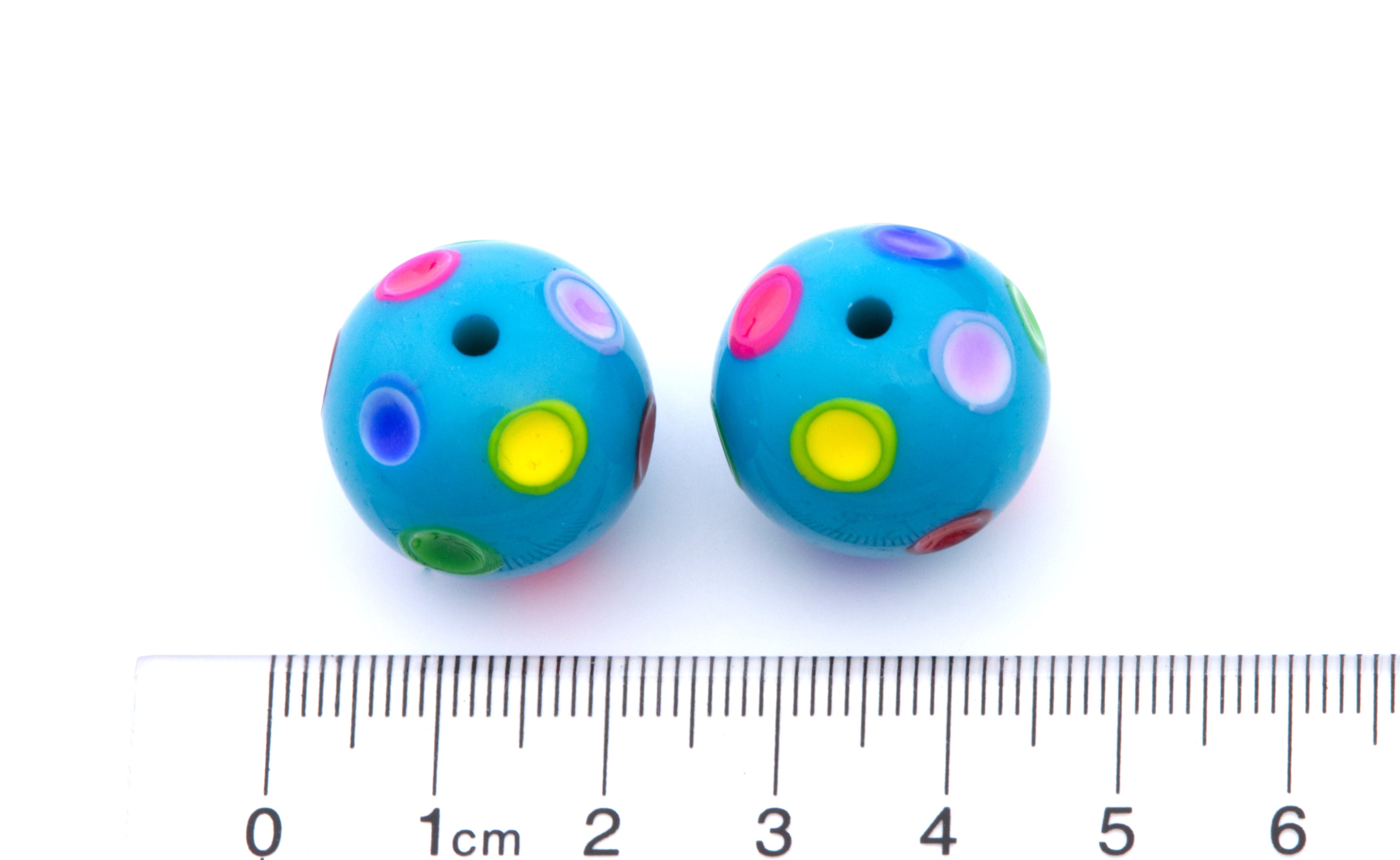 Two small blue round beads with colorful printed polka dots, shown next to a centimeter ruler for scale on a white background.