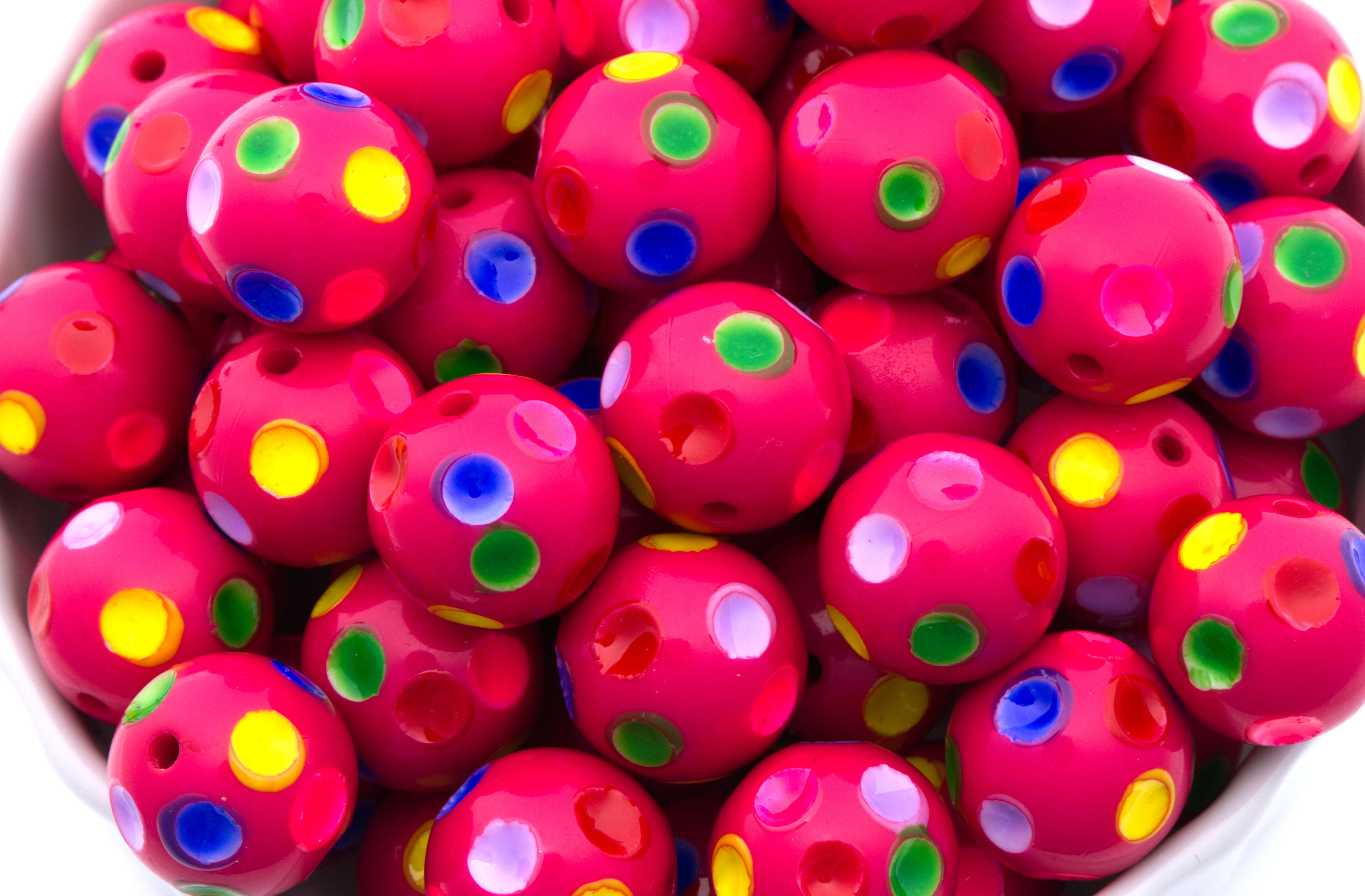 A close-up of many pink beads with colorful printed polka dots in yellow, green, blue, and white, piled together in a white container.