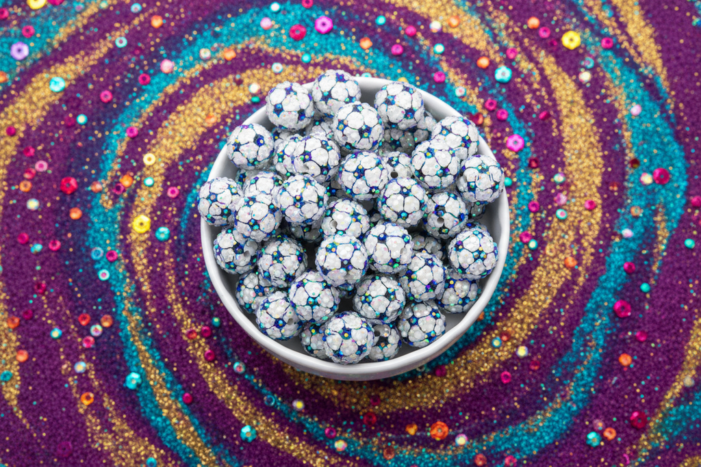 A white bowl filled with rhinestone ab soccer ball beads placed on a colorful glittery purple background with concentric rings of gold, teal, and pink sparkle.