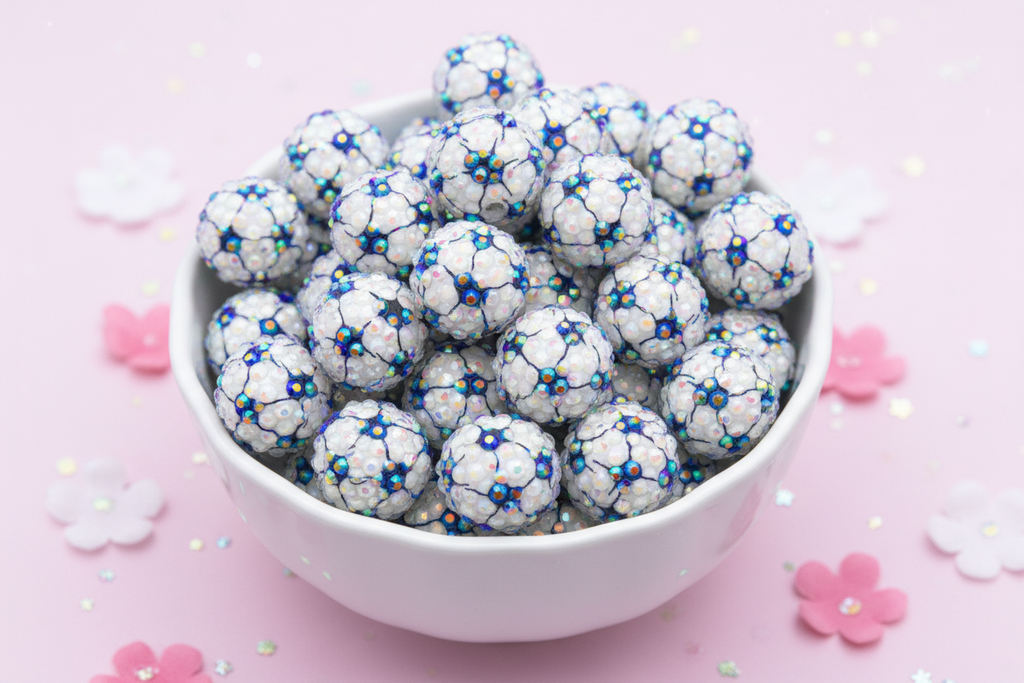 A white ceramic bowl filled with round rhinestone AB soccer ball beads, set against a light pink background with scattered decorative flower petals and glitter.