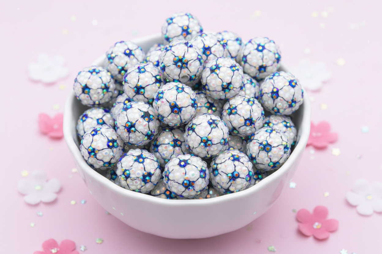 A white ceramic bowl filled with round rhinestone AB soccer ball beads, set against a light pink background with scattered decorative flower petals and glitter.