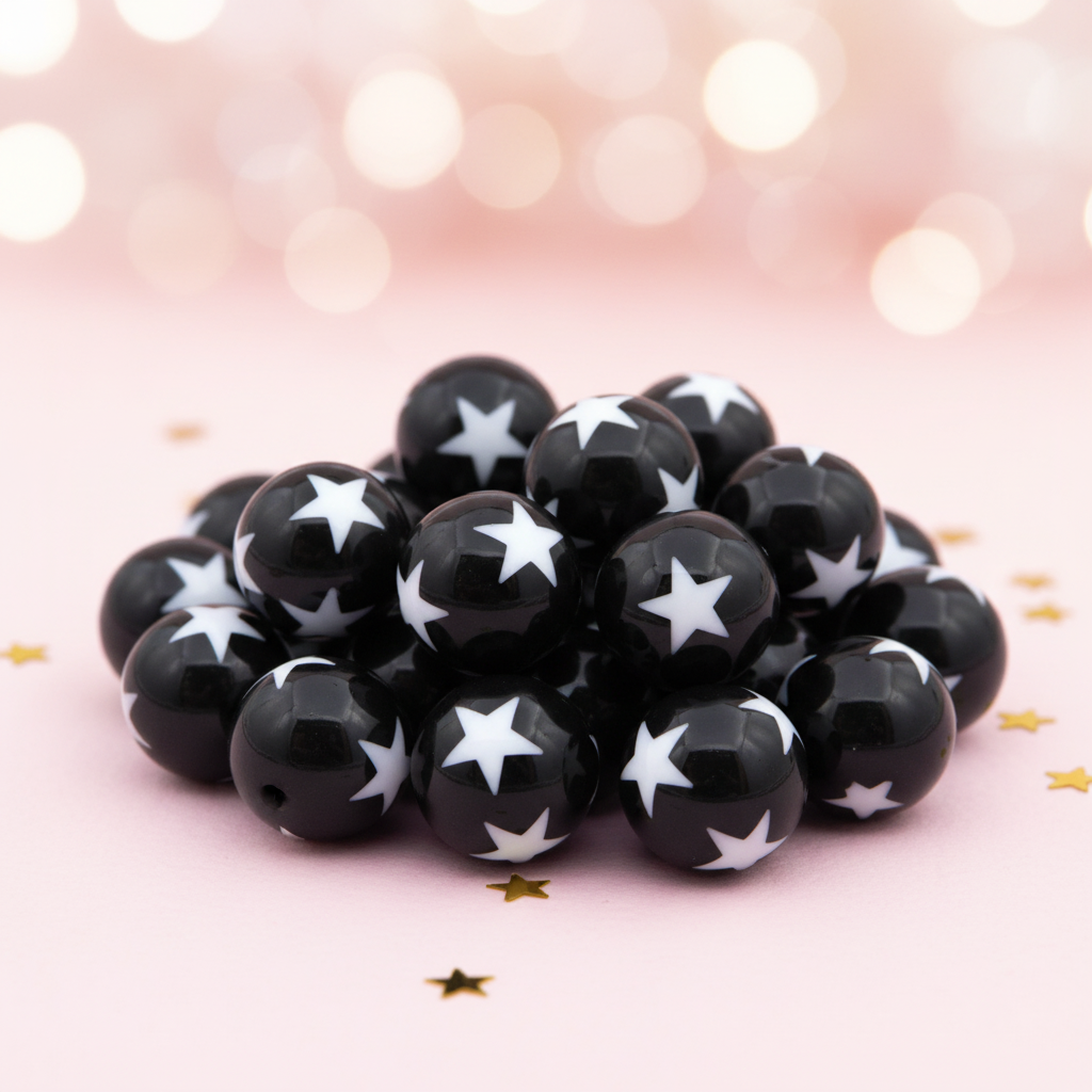 A pile of  black round beads with white star patterns, arranged on a soft pink background with gold star confetti and a blurred bokeh light background.