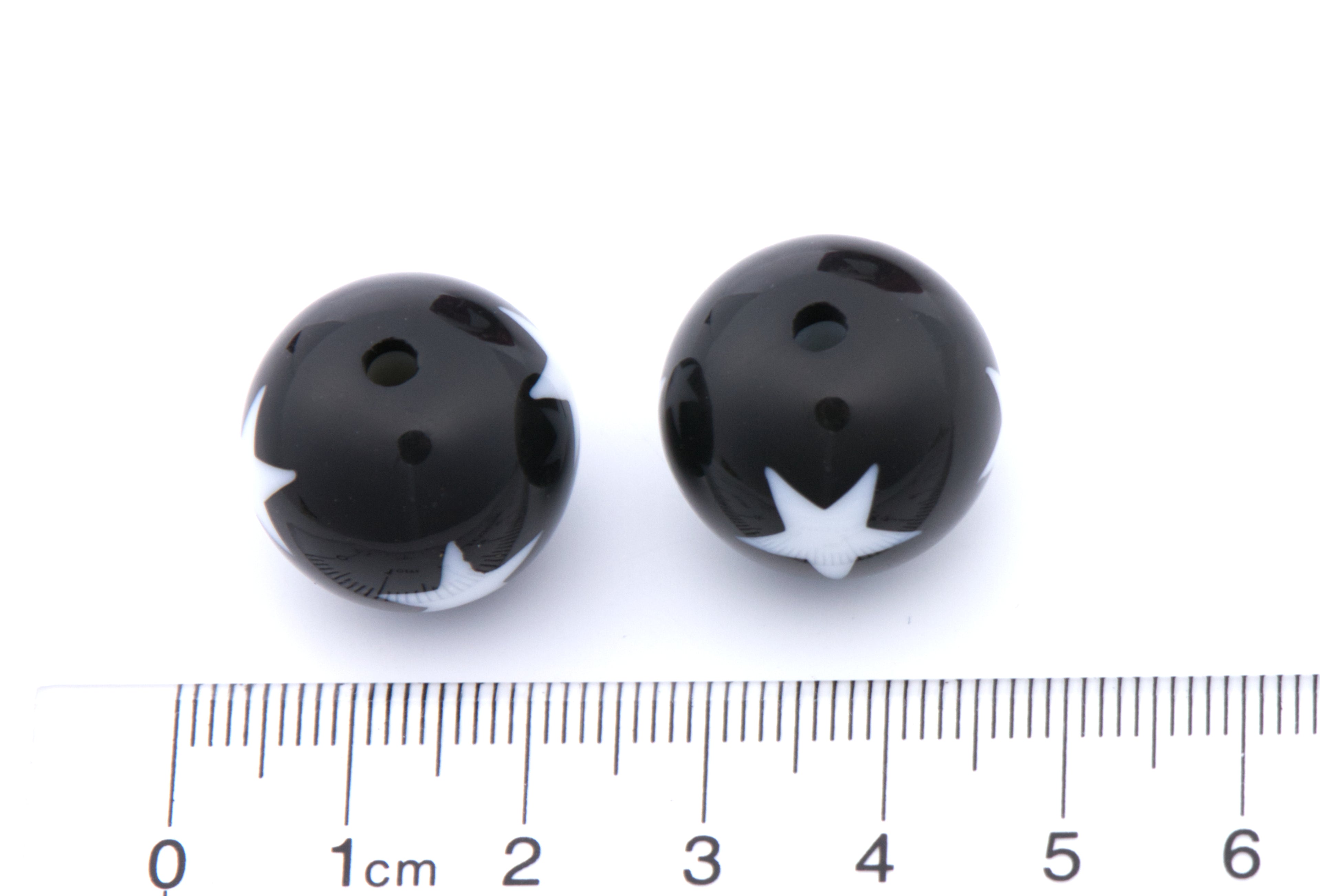 Two small black round beads with white star print, shown next to a cm ruler for scale on a white background.