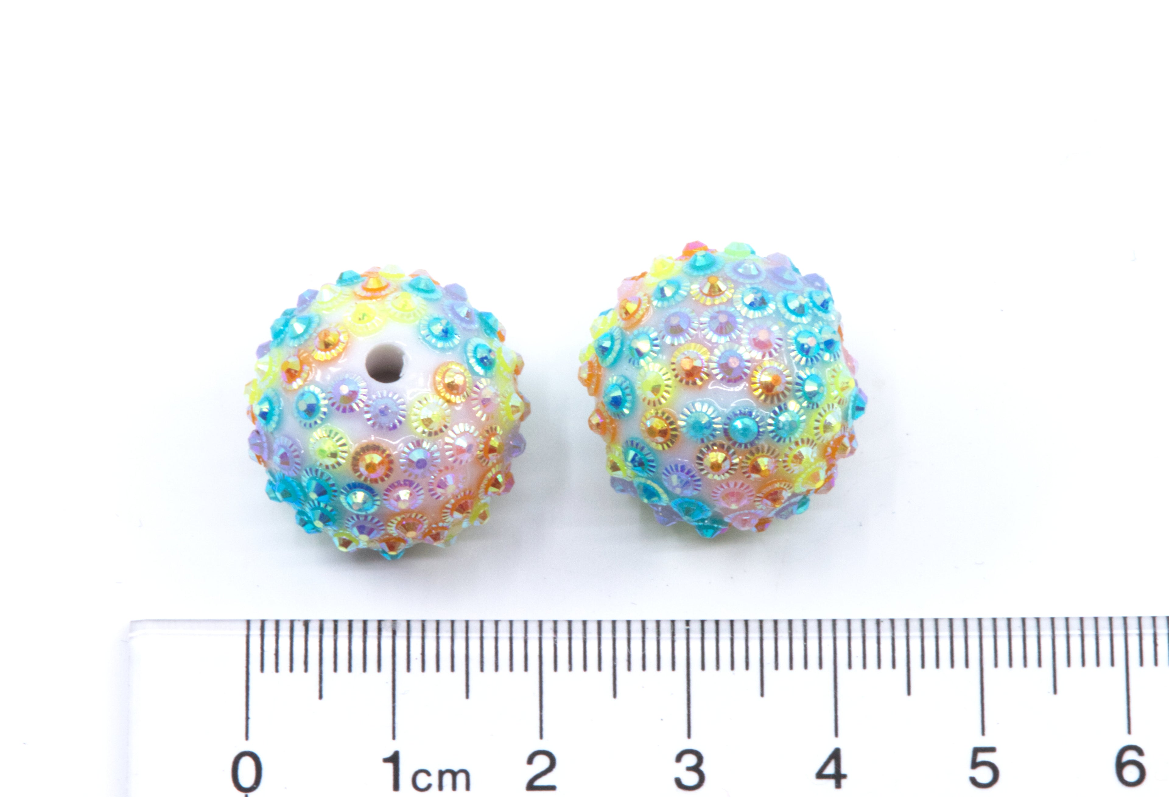 Two Rainbow rhinestone beads with a ruler for scale on a white background