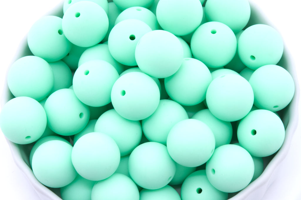 A close-up of many matte mint-green round beads, piled inside a white bowl against a white background.