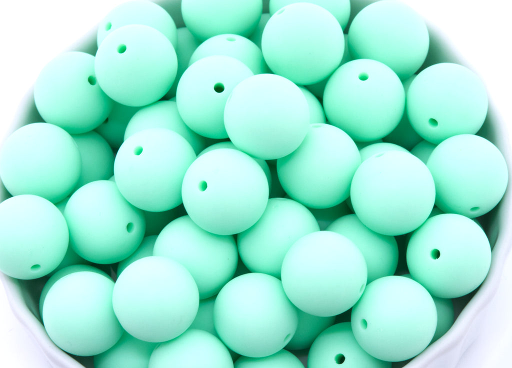 A close-up of many matte mint-green round beads, piled inside a white bowl against a white background.